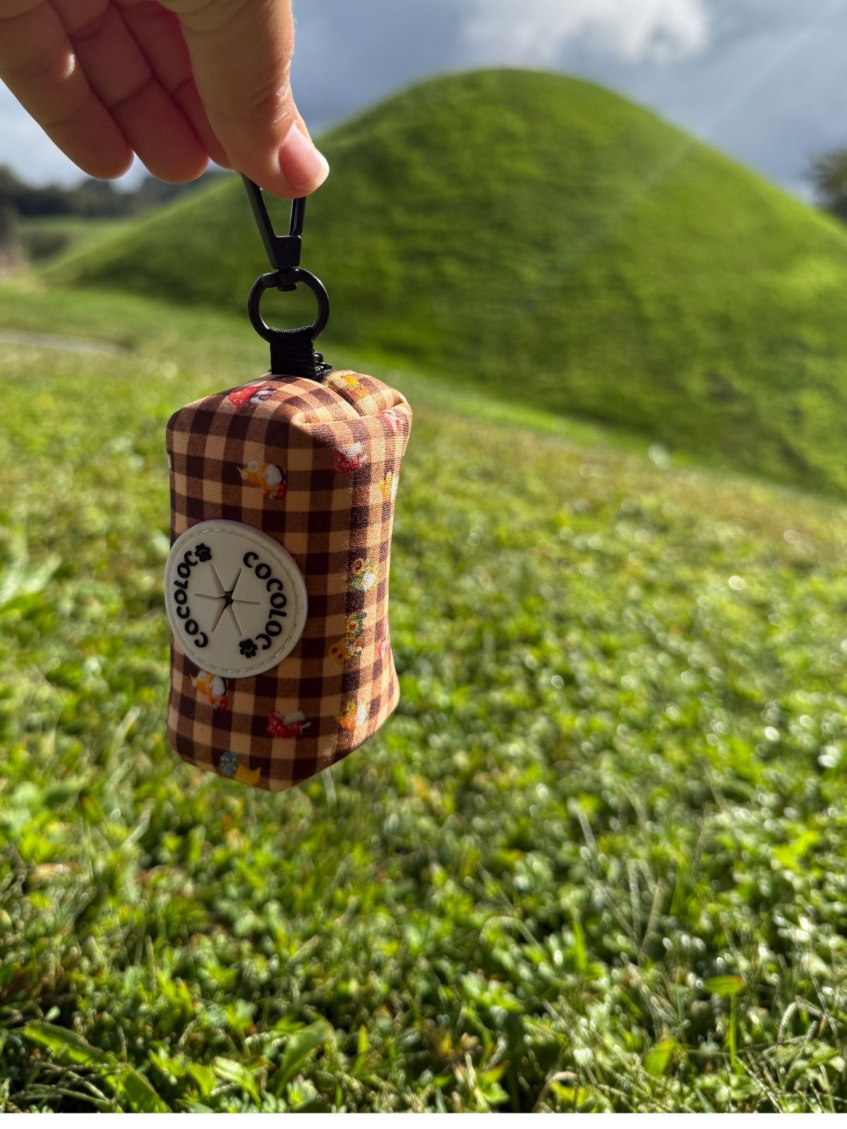 Porta bolsas Trasno colgado de su mosquetón con el logo COCOLOCO sobre un paisaje verde